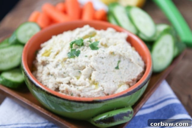 A close-up shot of creamy Lemony Artichoke Hummus in a white bowl, garnished with fresh herbs, ready to be enjoyed with a selection of vibrant raw vegetables.