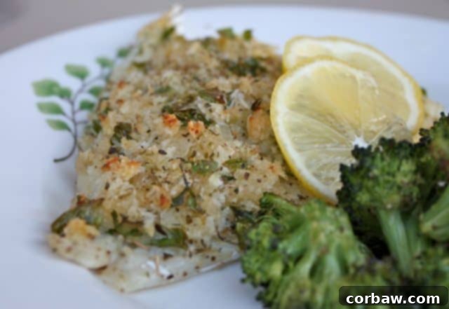 Baked breaded and herbed fish on a white plate, served with a side of roasted broccoli and a lemon wedge.