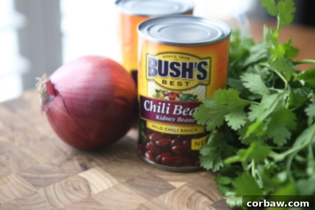 Chipotle Chicken Chili wooden cutting board with a red onion, two cans of Bush's chili bean kidney beans, and bunch of cilantro