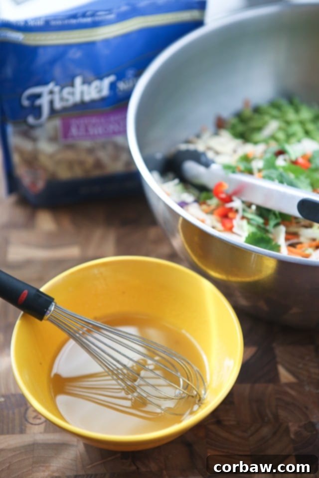 Asian Almond Slaw Salad A small yellow bowl holds the vibrant Asian dressing being whisked, positioned beside a larger blue bowl brimming with the colorful slaw ingredients and a package of Fisher sliced almonds, ready to be combined.