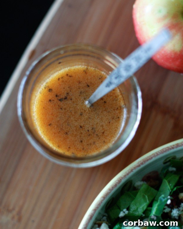 A top-down view of luscious maple vinaigrette in a clear glass jar, accompanied by a small spoon, ready to drizzle over a fresh salad. The dressing boasts a beautiful amber hue, indicative of its sweet and smoky flavor.