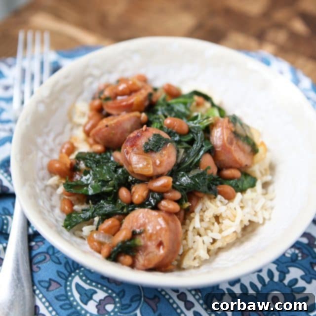 A comforting bowl of chicken sausage, flavorful baked beans, and vibrant kale served atop a bed of fluffy white rice, placed on a placemat with a fork.