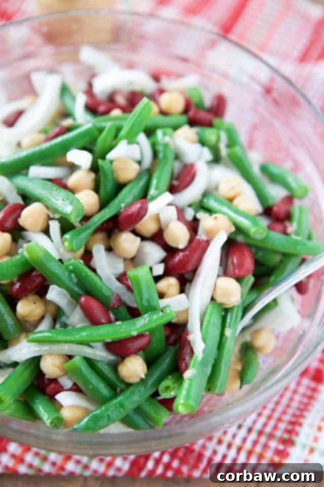 A vibrant Three Bean Salad featuring green beans, garbanzo beans, kidney beans, and thinly sliced Vidalia onions, tossed in a light dressing and presented in a clear glass bowl, perfect for a summer BBQ.