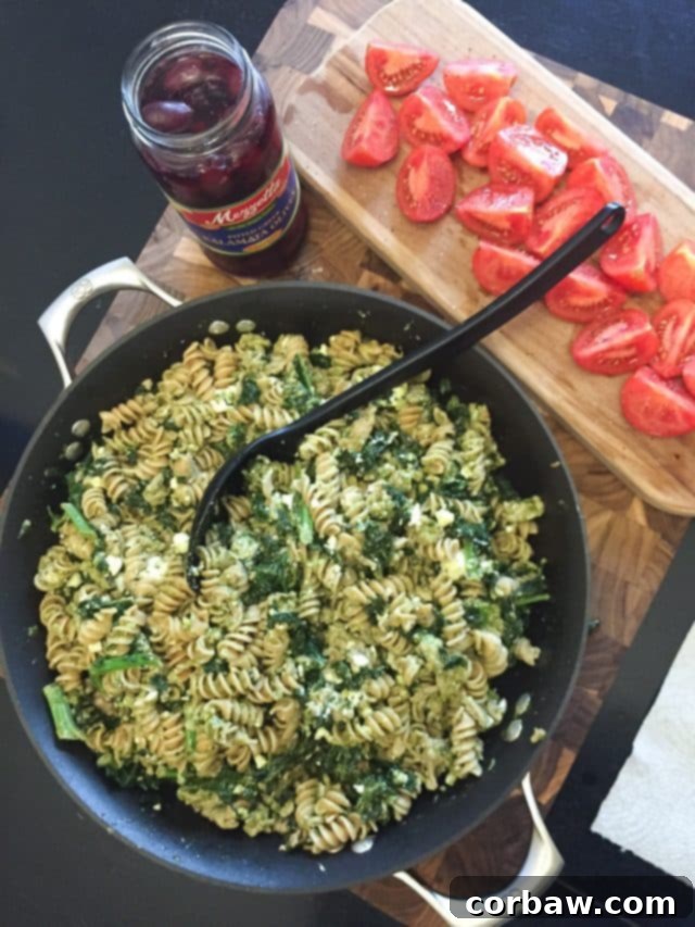A vibrant, large skillet brimming with healthy pesto pasta, savory ground turkey, and fresh kale, resting on a table alongside bright quartered tomatoes and Kalamata olives.