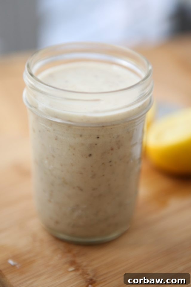 A beautiful mason jar brimming with the freshly prepared Parmesan Walnut Lemon Vinaigrette, elegantly displayed on a wooden cutting board with a halved lemon in the soft-focus background. Showcasing the final, creamy dressing for the Escarole Caesar Salad.