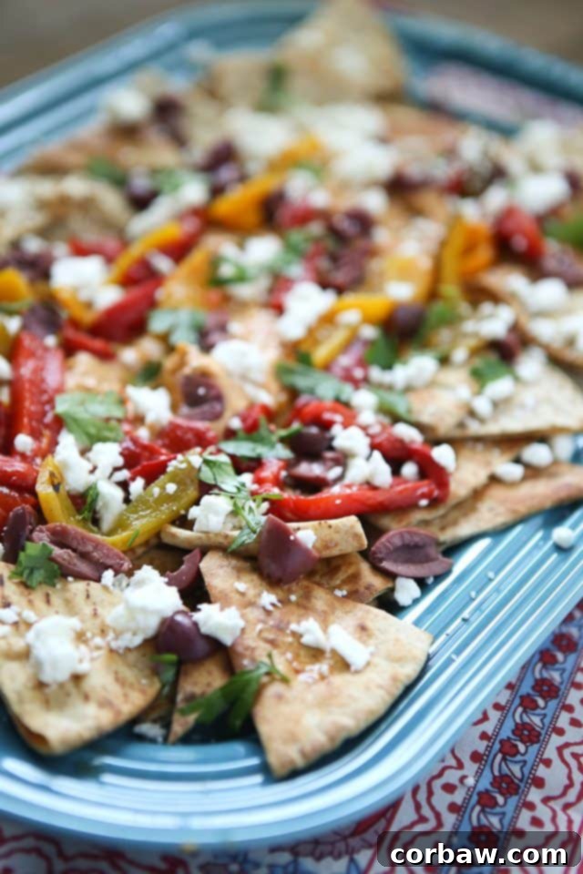 Mediterranean Nachos with Roasted Red Pepper Hummus: A Flavorful and Easy Appetizer Recipe A vibrant platter of Mediterranean Nachos, featuring baked pita chips generously topped with creamy Sabra Roasted Red Pepper Hummus, colorful roasted peppers, black olives, crumbled feta cheese, and fresh green herbs. This easy and delicious appetizer is perfect for any gathering or an unofficial meal.