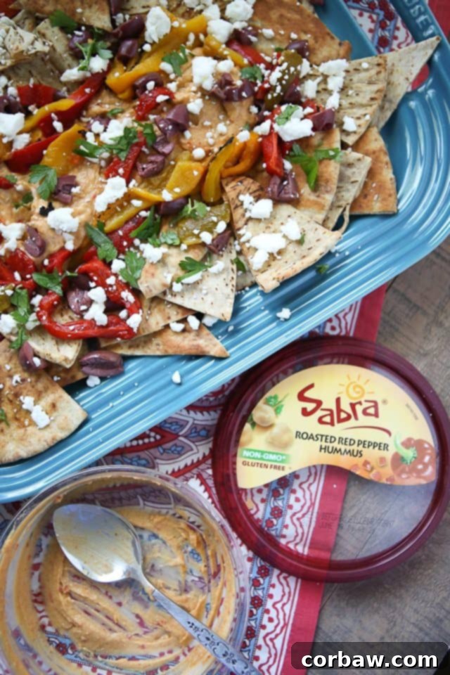 Easy Mediterranean Nachos with Sabra Roasted Red Pepper Hummus Close-up shot of delicious Mediterranean Nachos, piled high with baked pita chips, a generous dollop of Sabra Roasted Red Pepper Hummus, colorful roasted peppers, Kalamata olives, crumbly feta cheese, and fresh green parsley and oregano. A perfect, easy vegetarian appetizer.