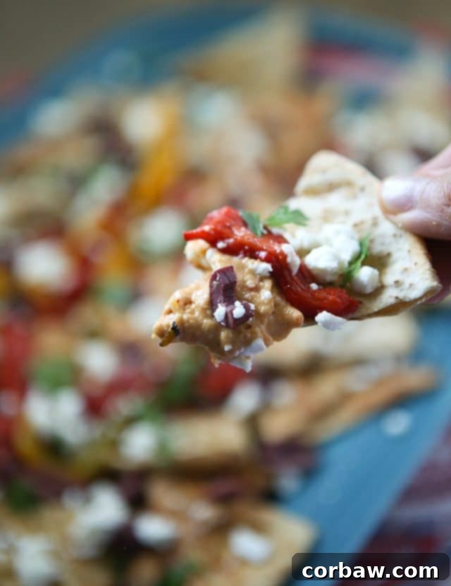 Mediterranean Nachos: Easy Recipe with Sabra Hummus and Fresh Toppings A top-down view of a vibrant serving plate filled with Mediterranean Nachos, featuring a generous scoop of Sabra Roasted Red Pepper Hummus surrounded by baked pita chips and topped with finely chopped roasted red peppers, kalamata olives, crumbled feta, and fresh green herbs. An ideal vegetarian appetizer or unofficial meal.