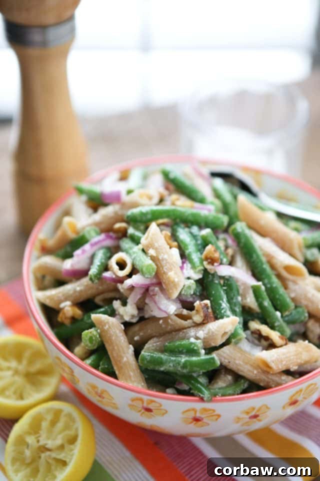Lemony Green Bean Pasta Salad with Walnuts: A refreshing, healthy, and easy vegetarian summer pasta salad. Lemony Green Bean Pasta Salad with Walnuts in a flowery bowl, garnished with fresh lemon, next to a pepper mill and salt bowl on a rustic table. Perfect for summer gatherings.