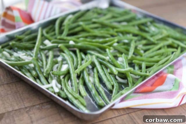 A sheet pan loaded with fresh green beans and thinly sliced onions, seasoned and ready for roasting.