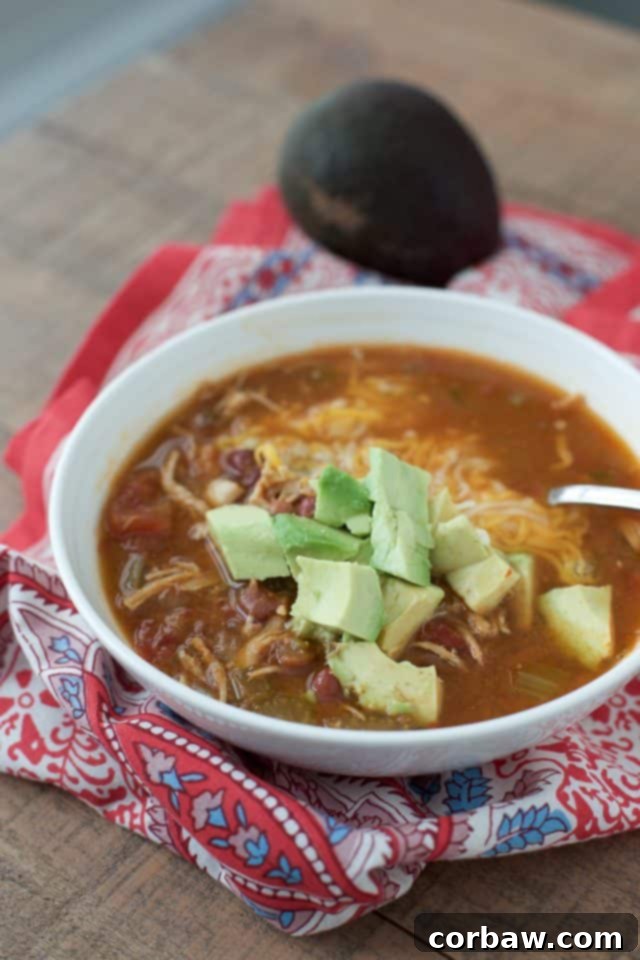 A white bowl of Instant Pot Mexican Chicken Soup, generously topped with diced avocado, rests on a red and blue napkin. A whole, ripe avocado sits in the background, hinting at the fresh, creamy garnish. This image highlights the comforting and healthy aspects of the soup, seasoned with chili, cumin, and smoked paprika, and filled with chicken, beans, and vegetables.