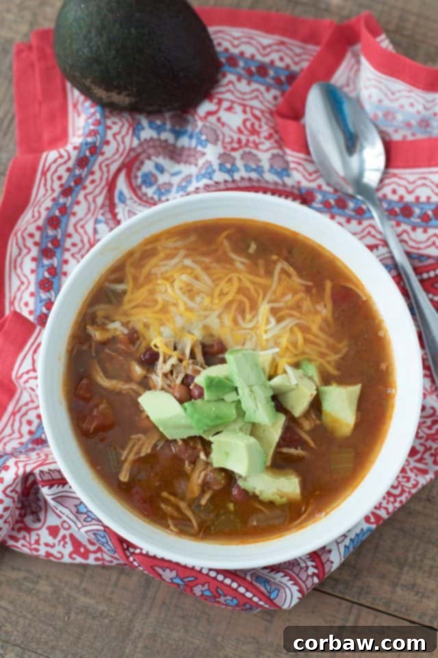 An inviting overhead shot of Instant Pot Mexican Chicken Soup. The soup is topped with a generous sprinkle of shredded cheese and perfectly diced avocado, with a spoon resting invitingly on a red and blue napkin. The image captures the rich texture and delicious toppings of this comforting, flavorful soup, emphasizing its healthy and easy preparation in the Instant Pot.