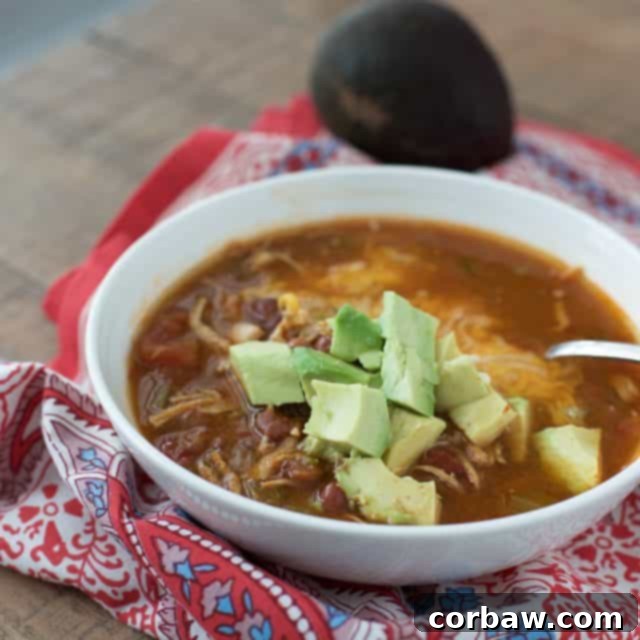 A beautifully presented white bowl of chili-flavored chicken soup, topped with generous cubes of fresh, diced avocado. The close-up shot emphasizes the vibrant colors and appealing texture of the soup, highlighting its rich flavor profile and healthy garnish. This image focuses on the delicious details of the Instant Pot Mexican Chicken Soup, perfect for a comforting meal.