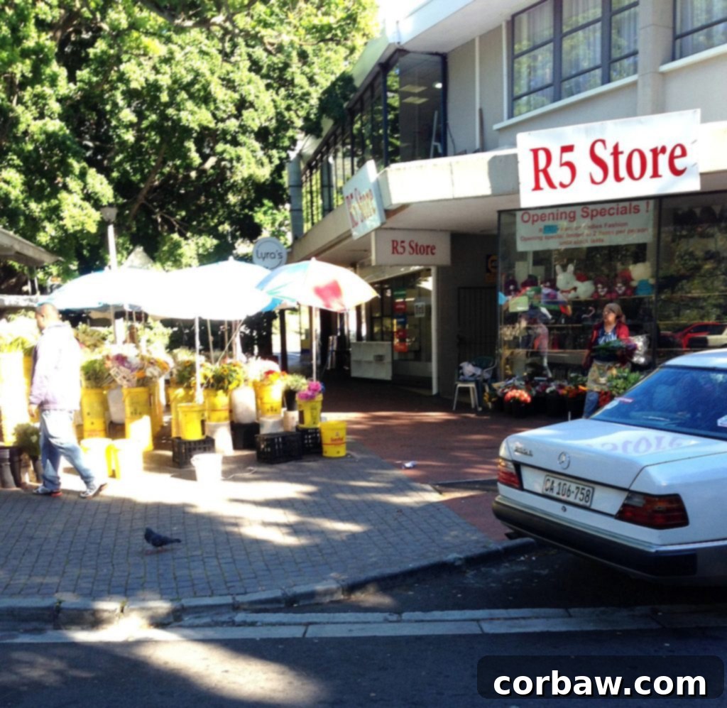 A street scene featuring a vibrant flower stall and a '5 Rand' store in Rondebosch.