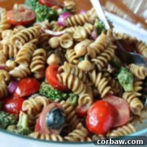 A bowl of vibrant pasta salad with cherry tomatoes, olives, red onions, and broccoli, garnished with fresh basil leaves.