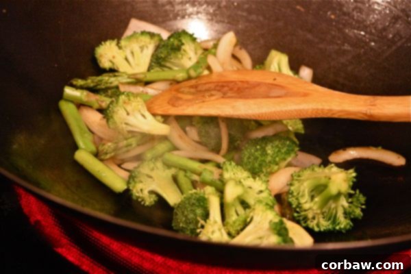Fresh asparagus, vibrant broccoli florets, and thinly sliced sweet onion being expertly stir-fried in a hot wok with a wooden spoon, showcasing the quick cooking process.