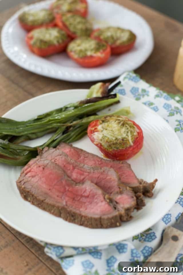 white plate with sliced london broil steak and green onion and roasted parmesan tomatoes in background