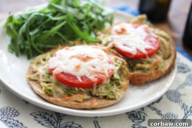 Close-up shot of a plate featuring two perfectly broiled English muffins, generously topped with shredded pesto chicken, fresh tomato slices, and gooey melted mozzarella cheese, served with a fresh side salad