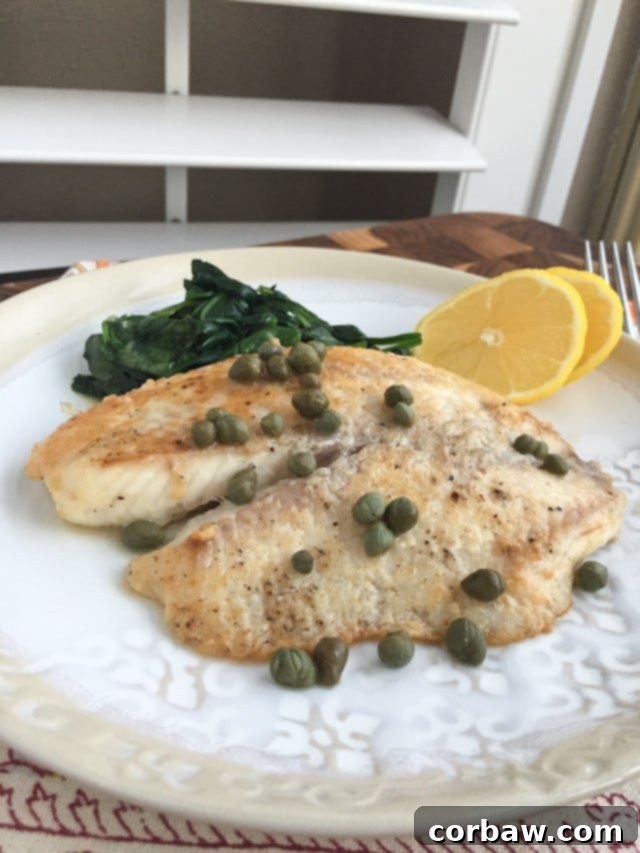 plate of white fish topped with capers with a side of cooked spinach and two lemon slices